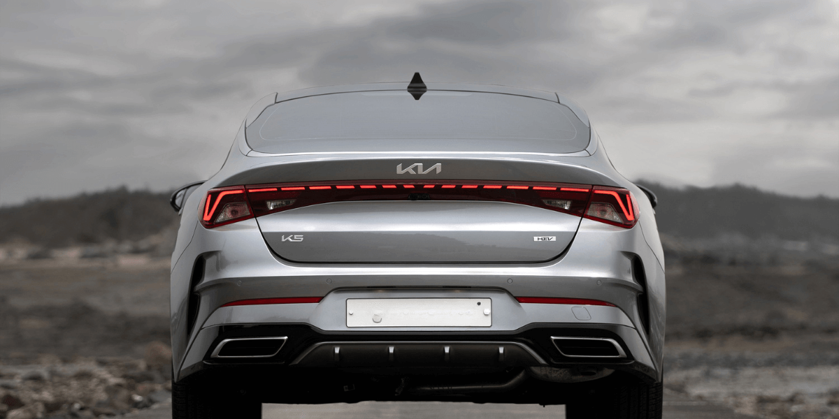 Rear view of a silver Kia K5 parked outdoors, highlighting the vehicle’s LED taillights and modern design against a cloudy landscape.